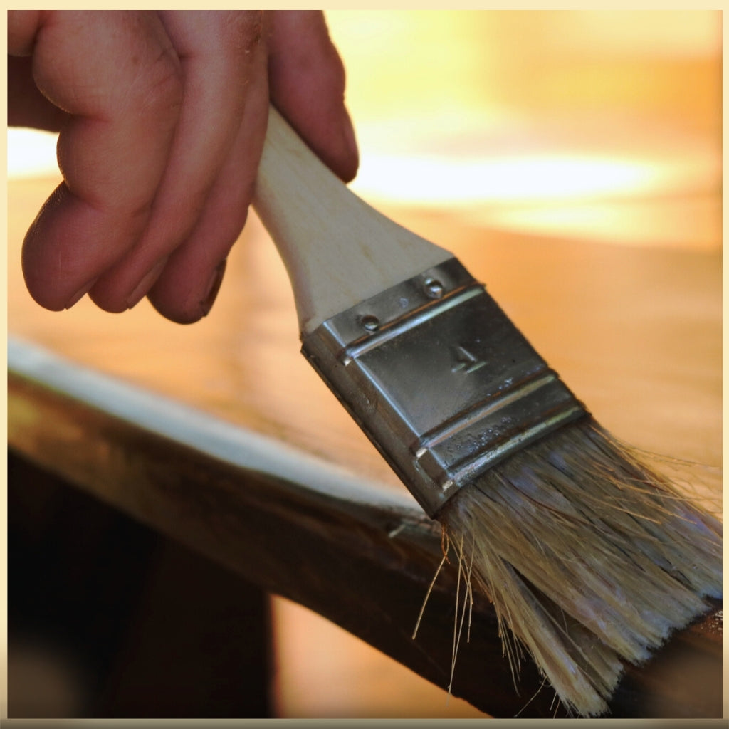 Hand holding paint brush applying products to a shiny wood table at sunset.