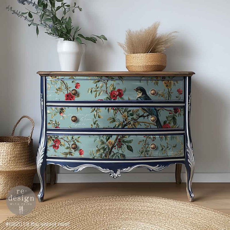 Vintage chest of drawers with floral painted design, dark blue trim, and silver detailing, alongside a woven basket and a white vase with greenery.