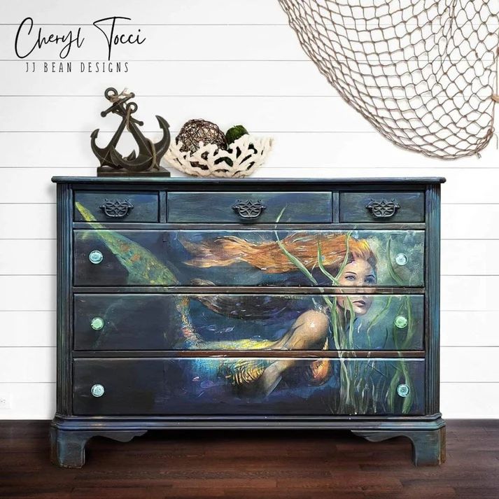A dark blue dresser with an artistic mermaid painting on the front, surrounded by decorative elements like an anchor and natural materials.