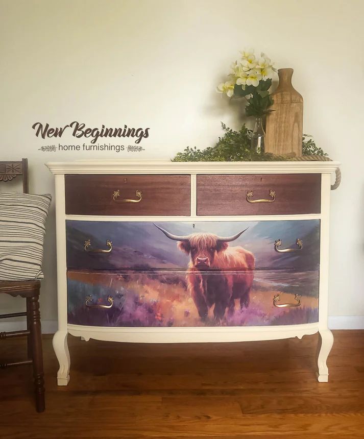 A vintage-style dresser painted white with wooden accents, featuring a colorful artwork of a Highland cow on the lower drawers, flanked by a vase of flowers and text reading "New Beginnings home furnishings."