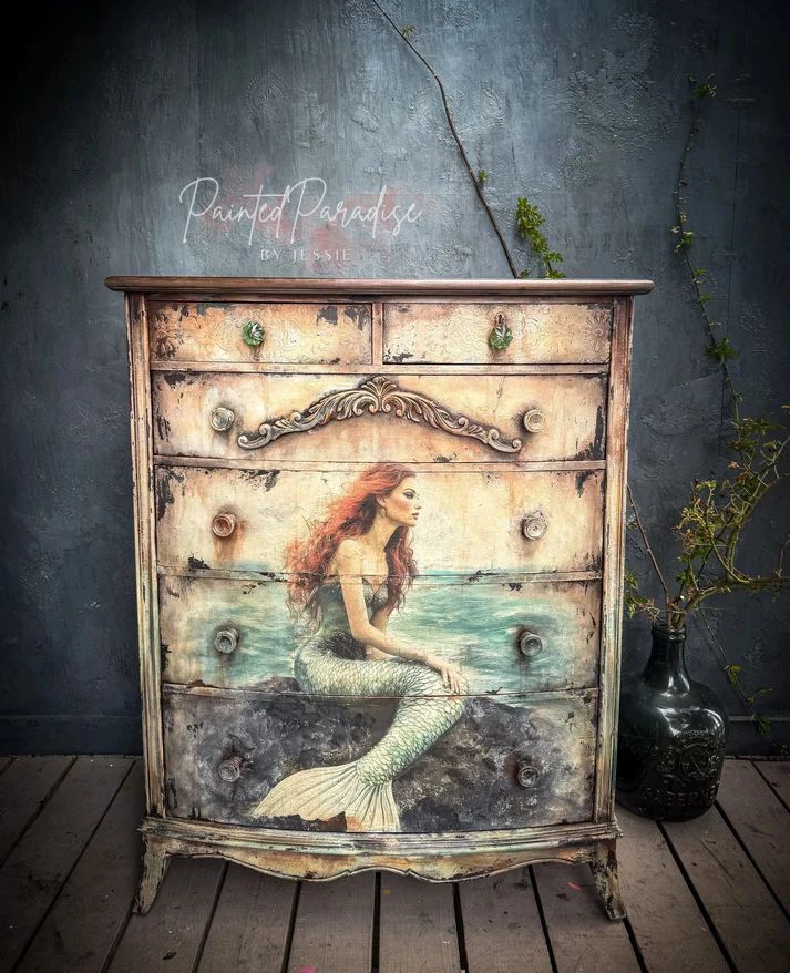 A vintage dresser painted with a mermaid design, featuring flowing red hair and a greenish tail, set against a textured gray wall